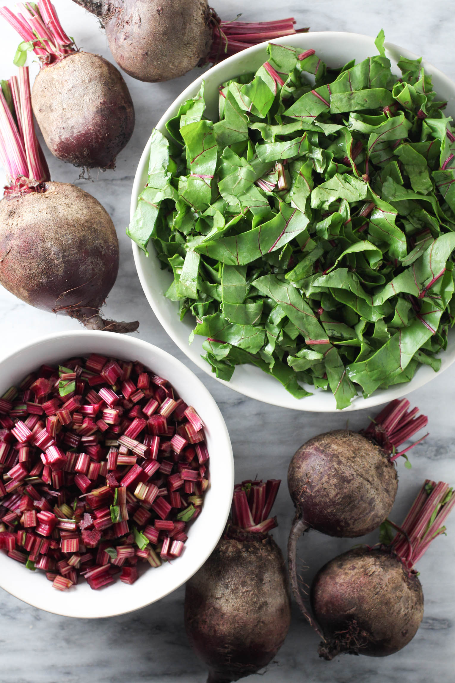 Chopped beet greens and stems in bowls and whole beets on marble background.