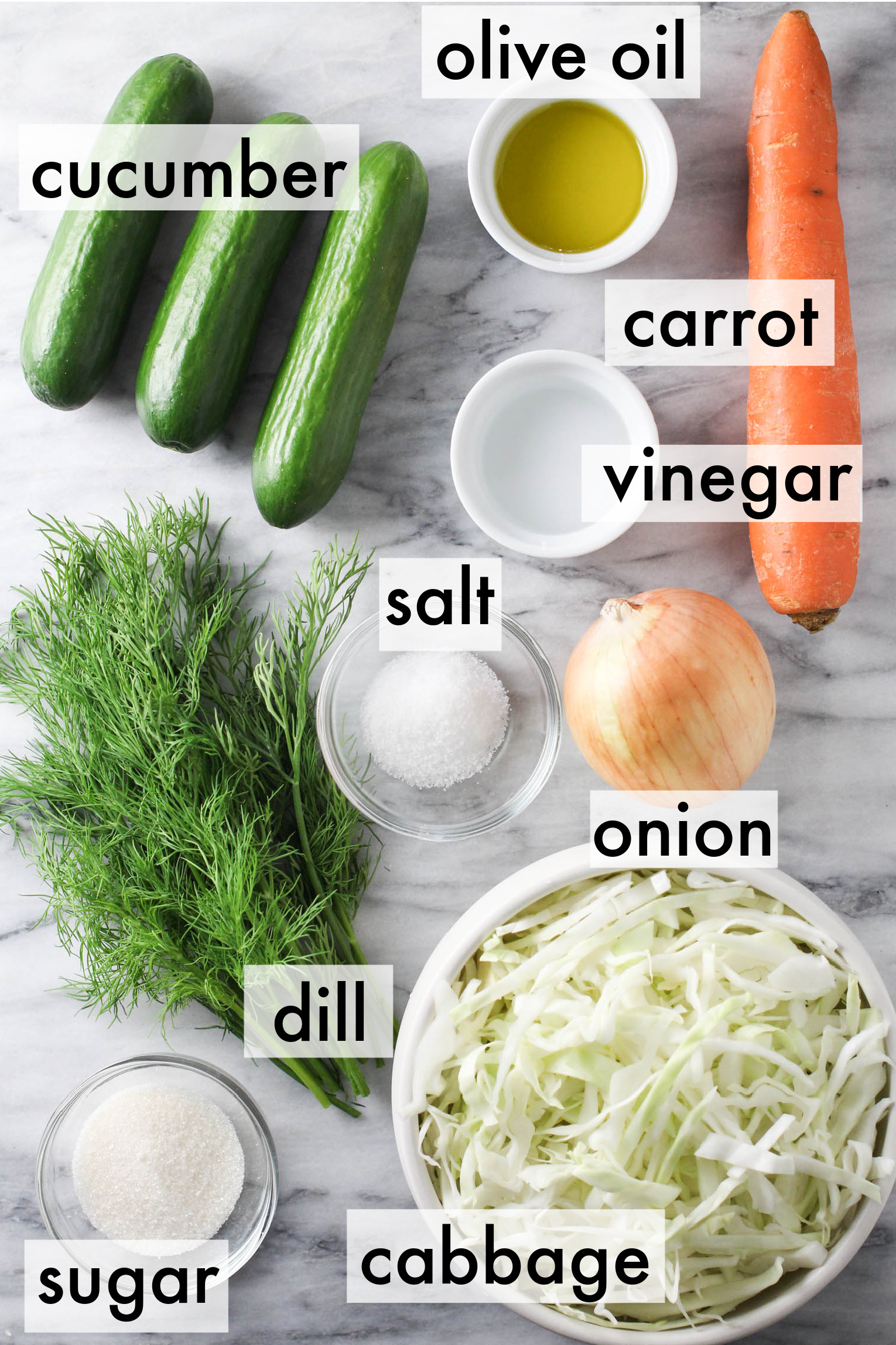 Cabbage cucumber salad ingredients displayed on marble background. The ingredients are labeled as follows: olive oil, cucumber, carrot, vinegar, salt, onion, dill, cabbage, sugar.