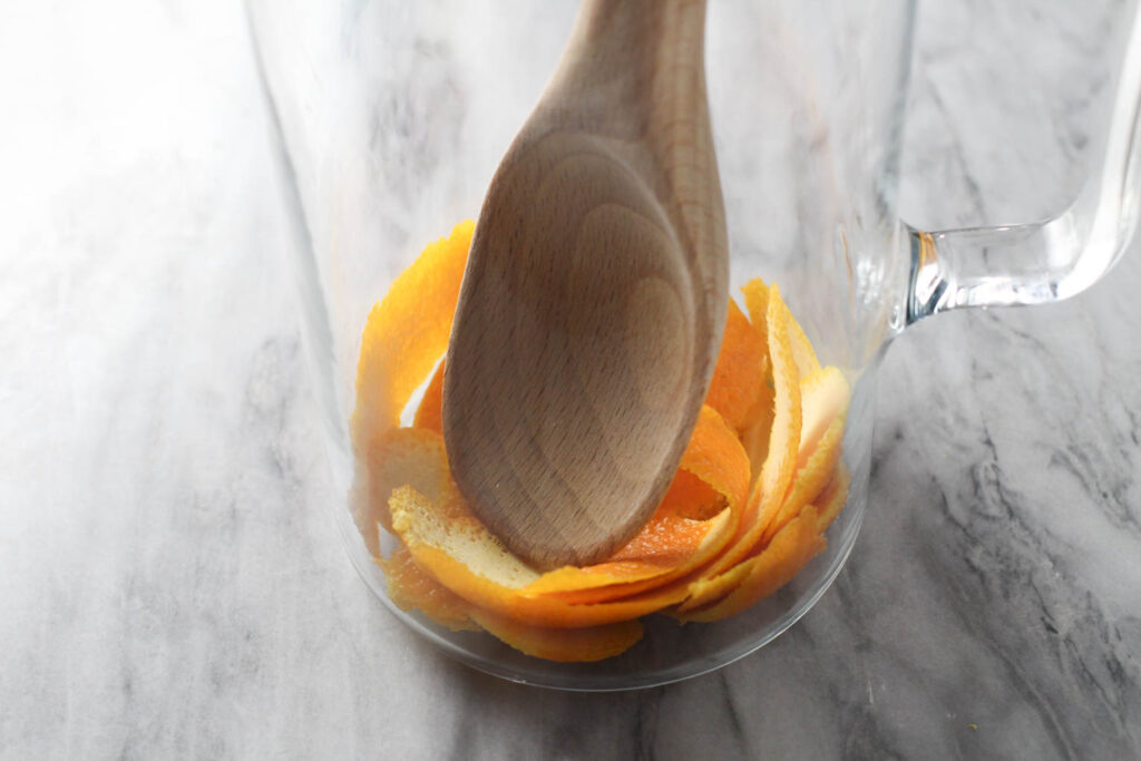 Orange peel on the bottom of a pitecher being muddled with a wooden spoon.