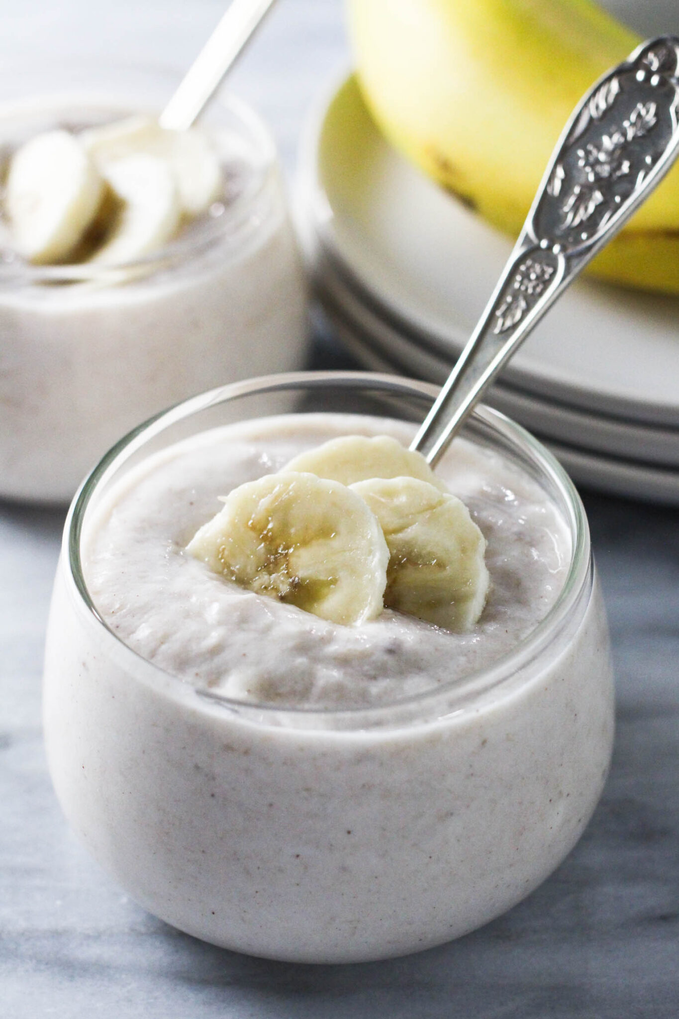 Banana yogurt in a glass bowl, garnished with banana slices. Plates and another bowl in the background.