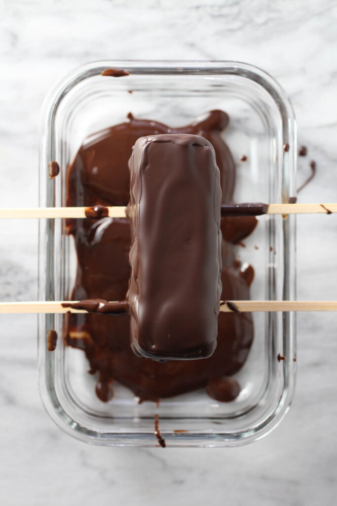 Frozen bar on top of skewers over a glass container, covered with chocolate.